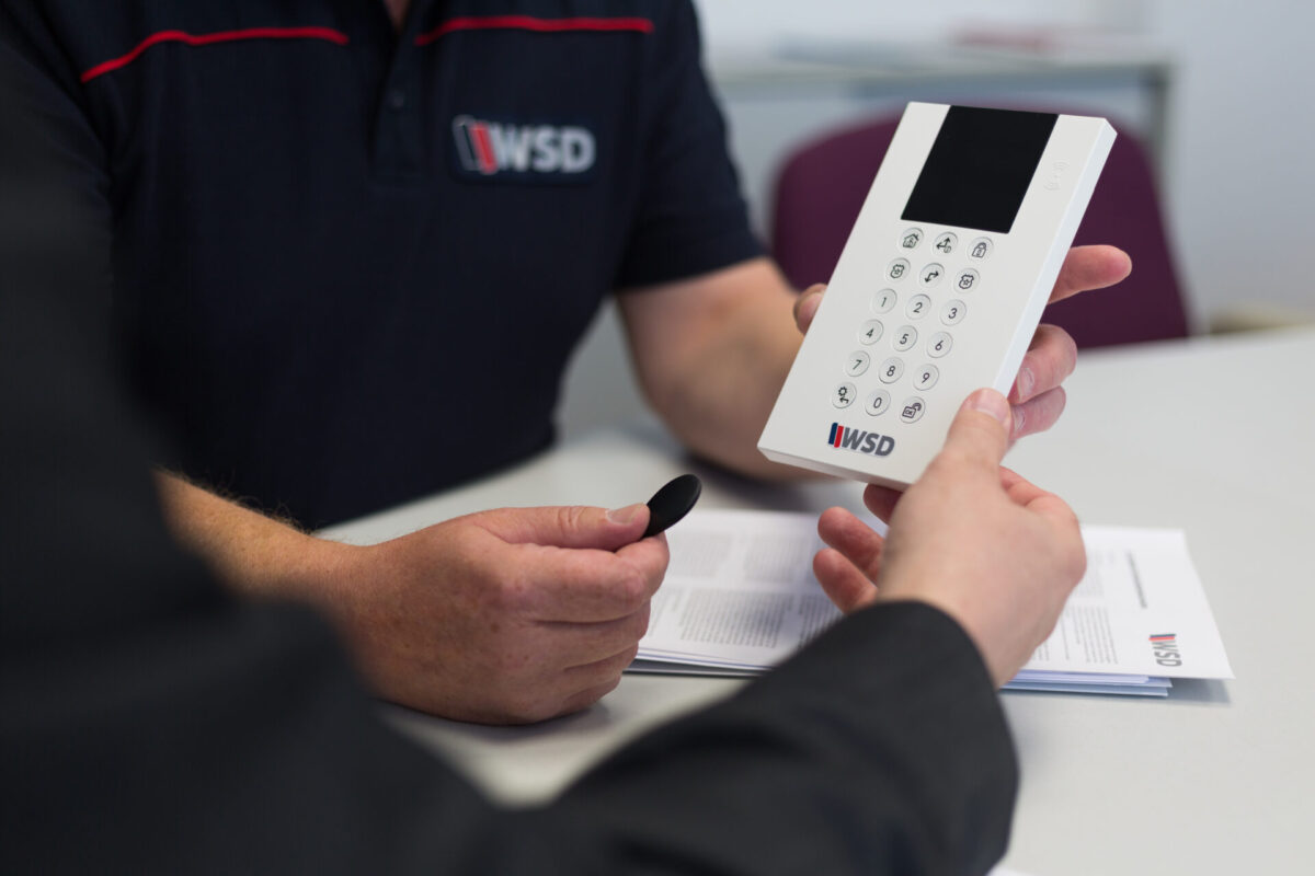 ChatGPT: Auf dem Bild sieht man zwei Personen, die gemeinsam ein Bedienteil einer Alarmanlage betrachten. Eine Person hält das weiße Gerät mit Tastatur und Display in der Hand, die andere hält einen schwarzen Transponder. Beide sitzen an einem Tisch, auf dem auch Unterlagen mit dem WSD-Logo liegen. Das Bild vermittelt eine professionelle Beratungssituation im Sicherheitsbereich.