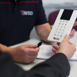 ChatGPT: Auf dem Bild sieht man zwei Personen, die gemeinsam ein Bedienteil einer Alarmanlage betrachten. Eine Person hält das weiße Gerät mit Tastatur und Display in der Hand, die andere hält einen schwarzen Transponder. Beide sitzen an einem Tisch, auf dem auch Unterlagen mit dem WSD-Logo liegen. Das Bild vermittelt eine professionelle Beratungssituation im Sicherheitsbereich.