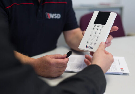 WSD-1029 ChatGPT: Auf dem Bild sieht man zwei Personen, die gemeinsam ein Bedienteil einer Alarmanlage betrachten. Eine Person hält das weiße Gerät mit Tastatur und Display in der Hand, die andere hält einen schwarzen Transponder. Beide sitzen an einem Tisch, auf dem auch Unterlagen mit dem WSD-Logo liegen. Das Bild vermittelt eine professionelle Beratungssituation im Sicherheitsbereich.